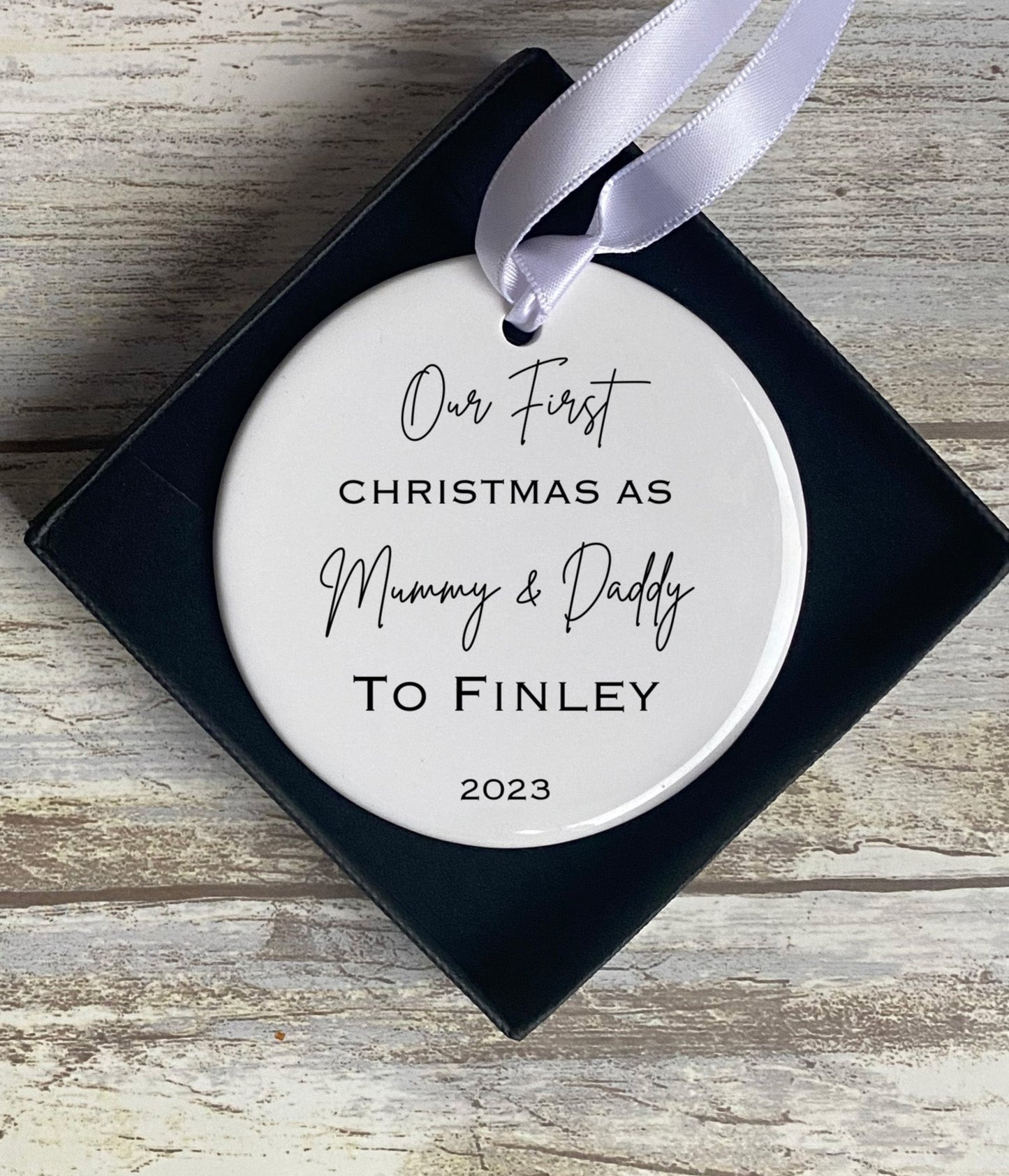Round ceramic Christmas ornament in a black gift box with white ribbon, featuring the message “Our First Christmas As Mummy & Daddy To Finley 2023” in elegant black script and serif fonts. Set against a rustic wooden background.