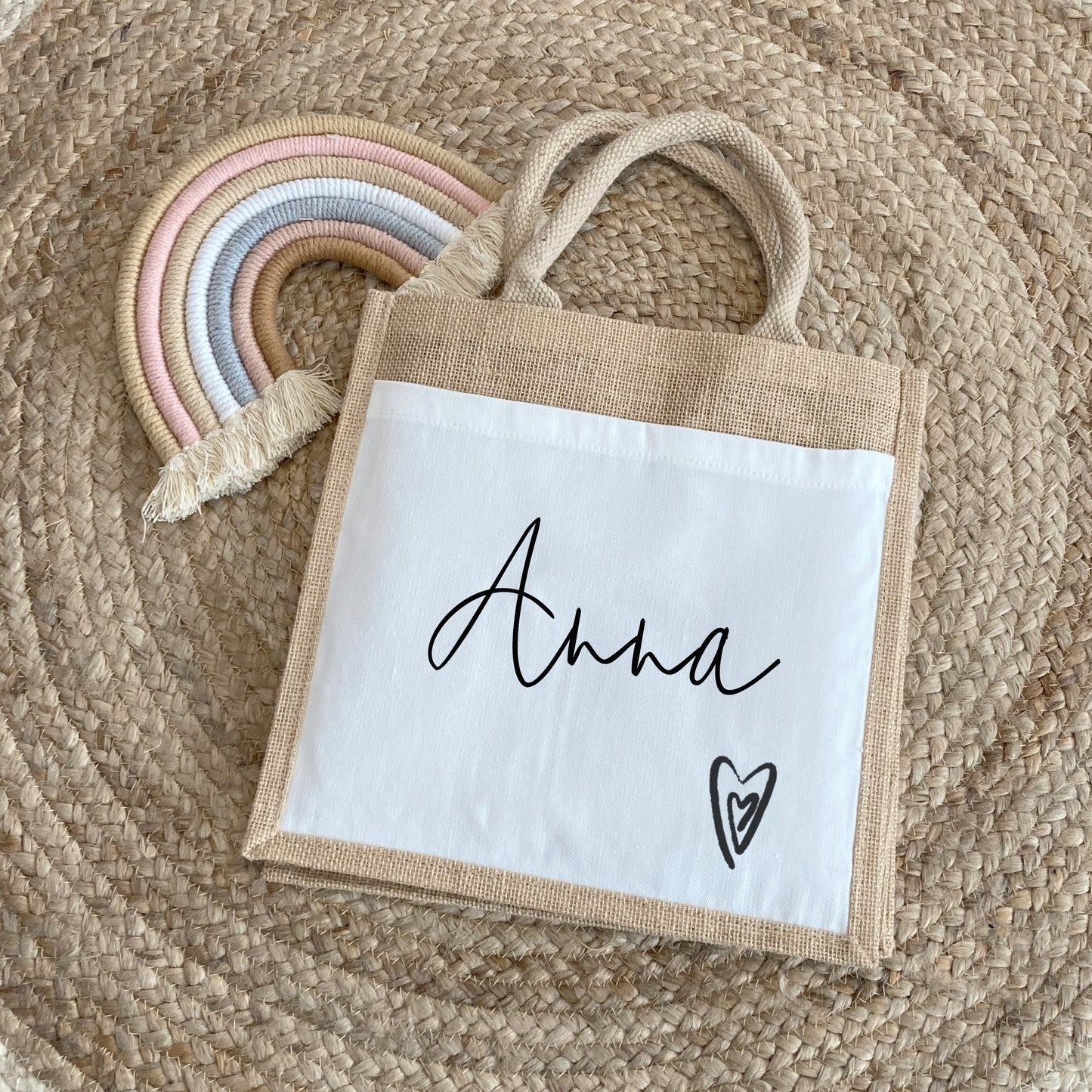 A personalized jute bag with a white canvas front panel printed with the name "Anna" in a graceful black script. A small black heart illustration appears at the bottom right. The bag rests on a round woven rug beside a pastel macrame rainbow.