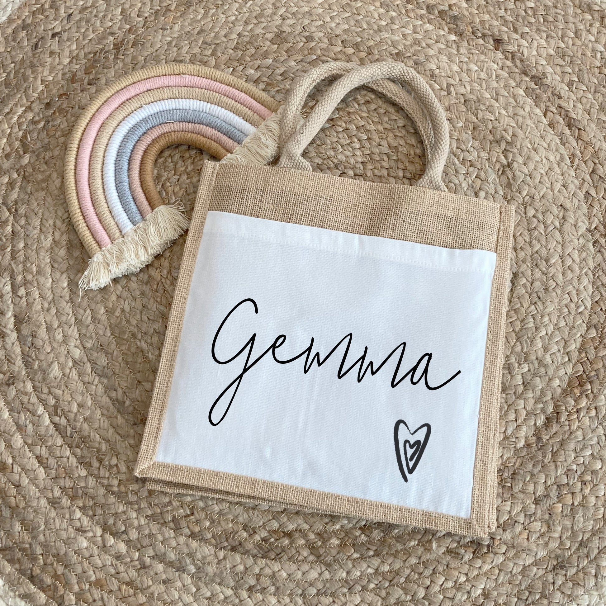 A natural jute bag with a white canvas front panel, personalized with the name "Gemma" in elegant black script. A small hand-drawn heart is printed near the bottom right corner. The bag is placed on a circular woven rug beside a pastel-colored macrame rainbow.