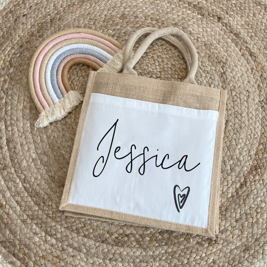 A personalized jute lunch bag with beige handles and trim, placed on a woven circular rug. The front white canvas panel displays the name “Jessica” in an elegant handwritten script font. Below the name, toward the bottom right corner, is a simple sketched heart outline with a smaller heart inside. A pastel rainbow rope decoration peeks out from the upper left corner, adding a soft touch to the scene.