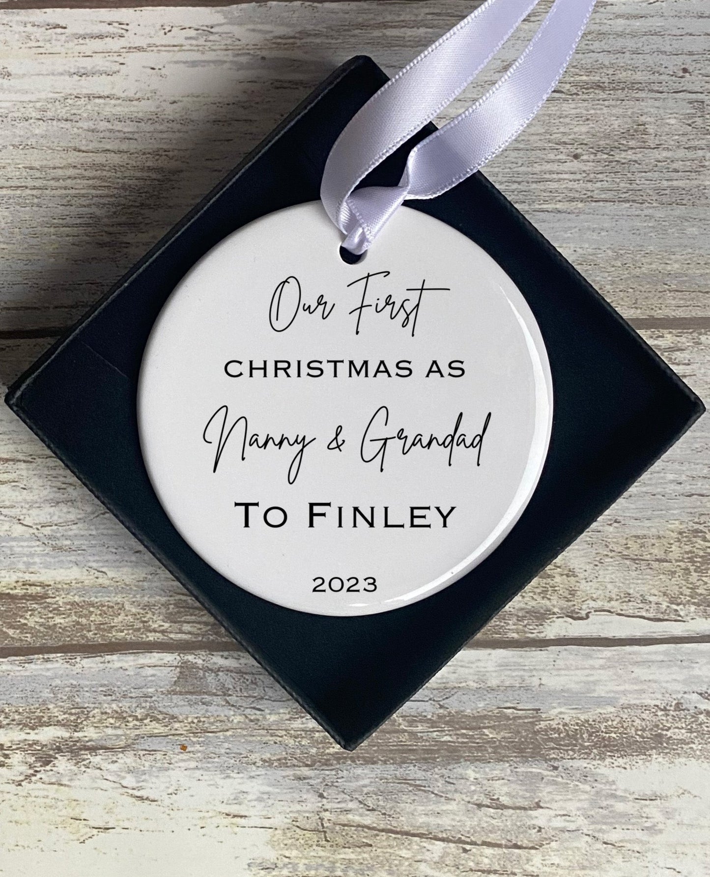 Ceramic round Christmas ornament with a white satin ribbon, displayed in a navy gift box. The ornament reads “Our First Christmas As Nanny & Grandad To Finley 2023” in mixed script and serif fonts. The background is a rustic wooden surface.