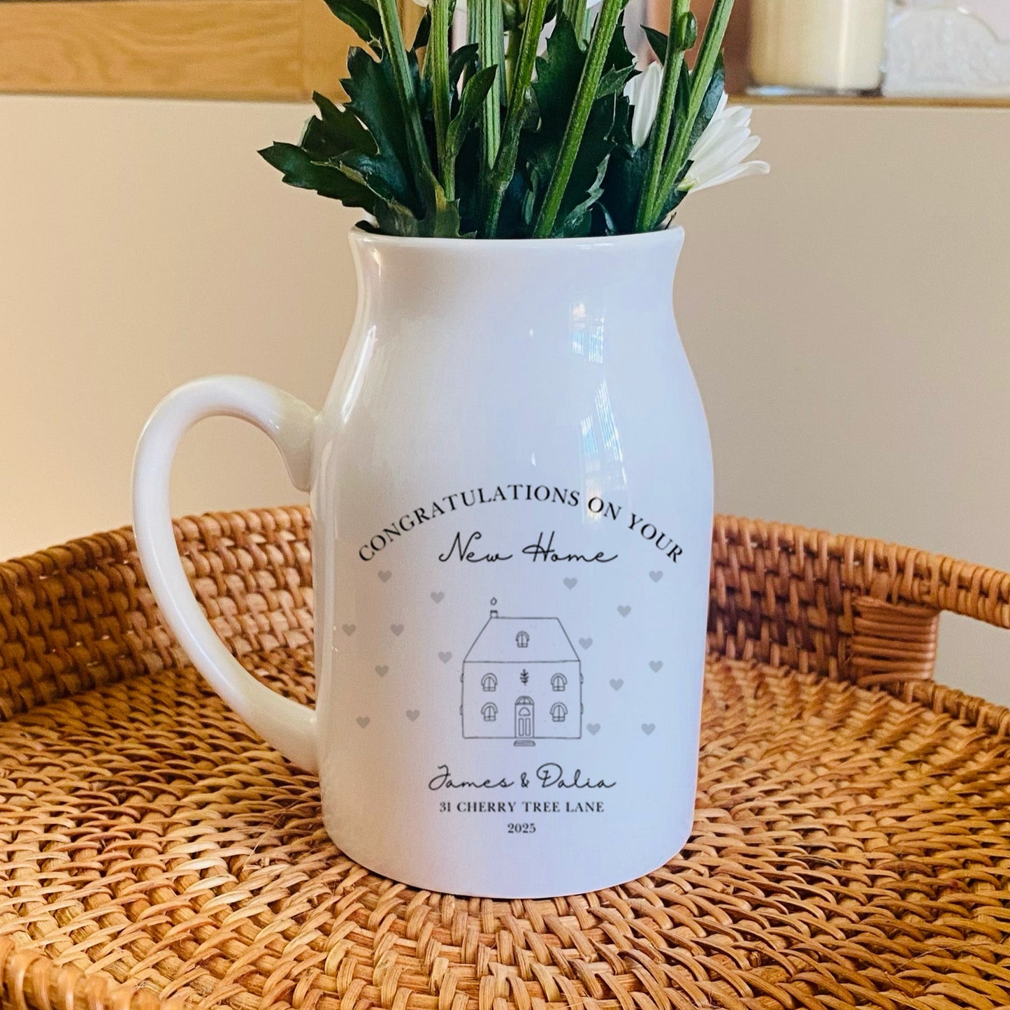 A personalised White pitcher vase with flowers on a woven tray in a home setting. The wording includes 'Congratulations on your new home'
