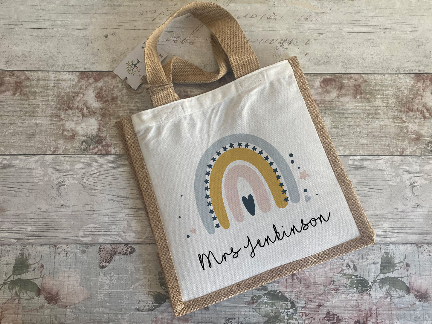 Personalised jute tote bag with a white canvas front panel featuring a pastel rainbow in mustard, grey, and pink tones, surrounded by small stars and dots. Below the rainbow is the name "Mrs Jenkinson" in a playful script font.