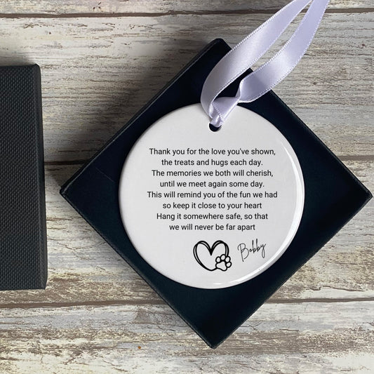 A round, white ceramic ornament with a printed poem that says: "Thank you for the love you've shown, the treats and hugs each day. The memories we both will cherish, until we meet again some day. This will remind you of the fun we had so keep it close to your heart. Hang it somewhere safe, so that we will never be far apart." At the bottom, there's a heart with a paw print design and the name "Bobby" in cursive. The ornament has a satin ribbon for hanging and is placed in a black gift box.