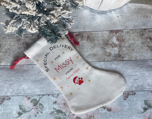 Personalised pet Christmas stocking with red drawstring, printed with 'Special Delivery for Missy from Santa Paws' and a red paw print, displayed on rustic wood background next to frosted tree