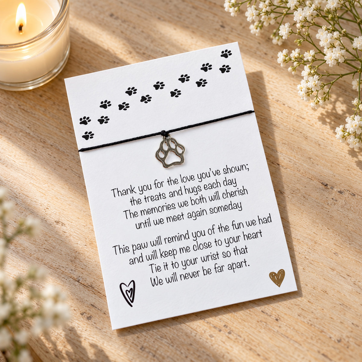 A pet memorial card with a black cord band and a paw print charm, displayed on a wooden surface with a lit candle and small white flowers.