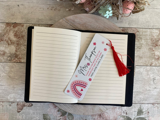 Open lined notebook with a white bookmark lying diagonally across the pages. The bookmark features the name 'Mrs Thompson' in script, a thank-you message from a student, red floral accents, a red rainbow illustration, and a red tassel. Set on a rustic floral wooden surface with a pastel egg and flower decoration at the top.