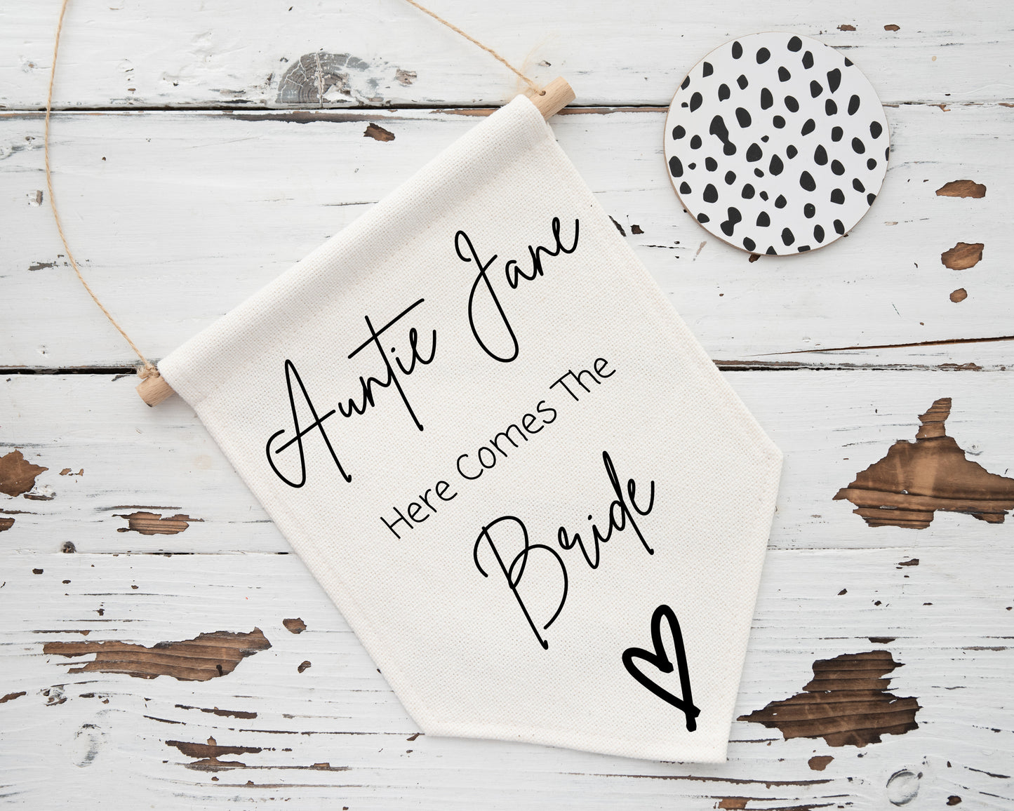 A cream-colored hanging linen flag placed on a distressed white wooden surface, featuring the text āAuntie Jane, Here Comes The Brideā in a mix of elegant cursive and clean sans-serif fonts. A small black heart illustration appears at the bottom right. A round black-and-white spotted coaster is also part of the scene.