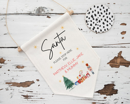 Personalised Christmas Santa stop here banner with names – festive linen hanging flag featuring gifts, Christmas tree, and nutcracker – rustic wooden background decor