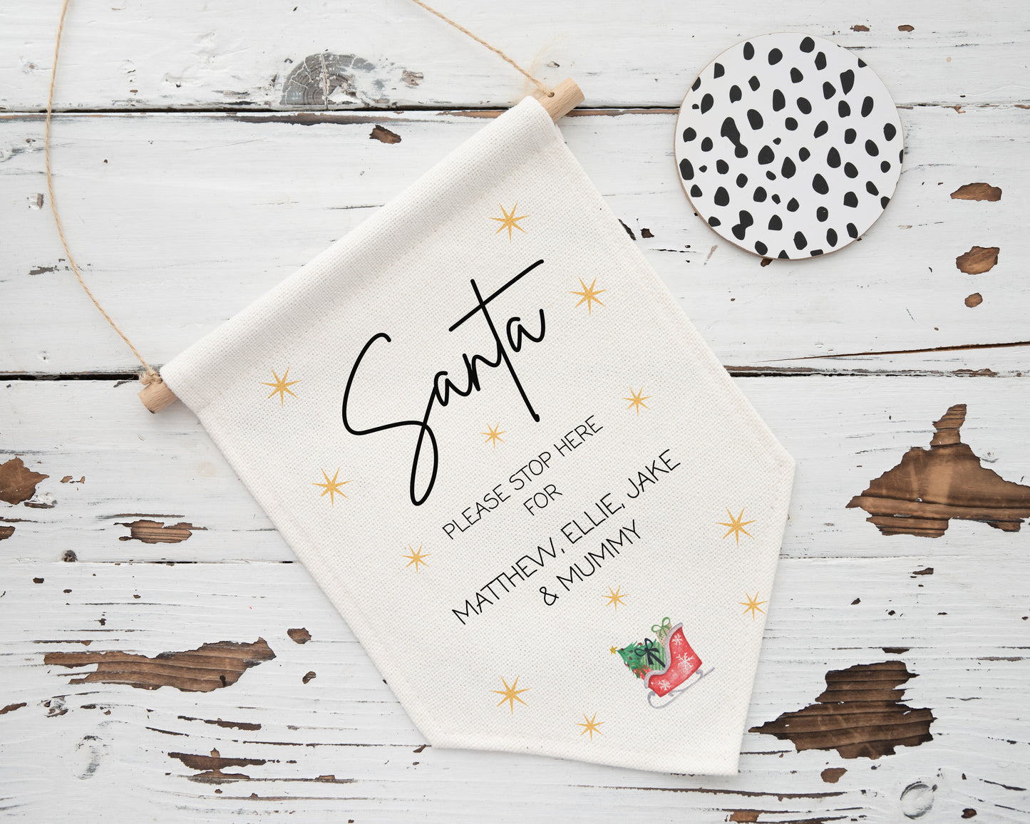 Personalised "Santa Please Stop Here" hanging flag with family names and festive sleigh design, displayed on rustic wooden background with black and white coaster