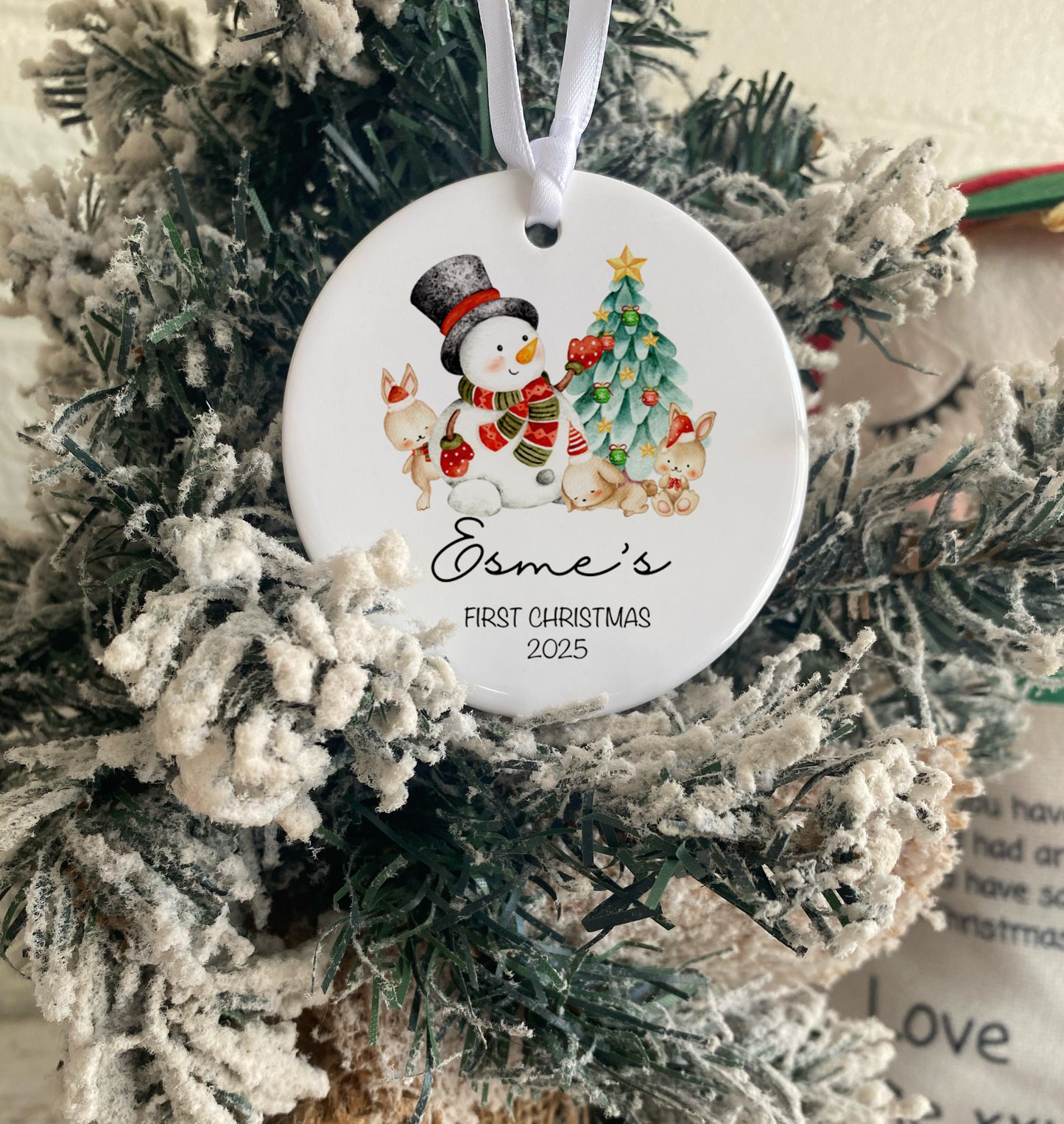 Decorative Christmas ornament with a snowman and text 'Esme's First Christmas 2025' on a tree.