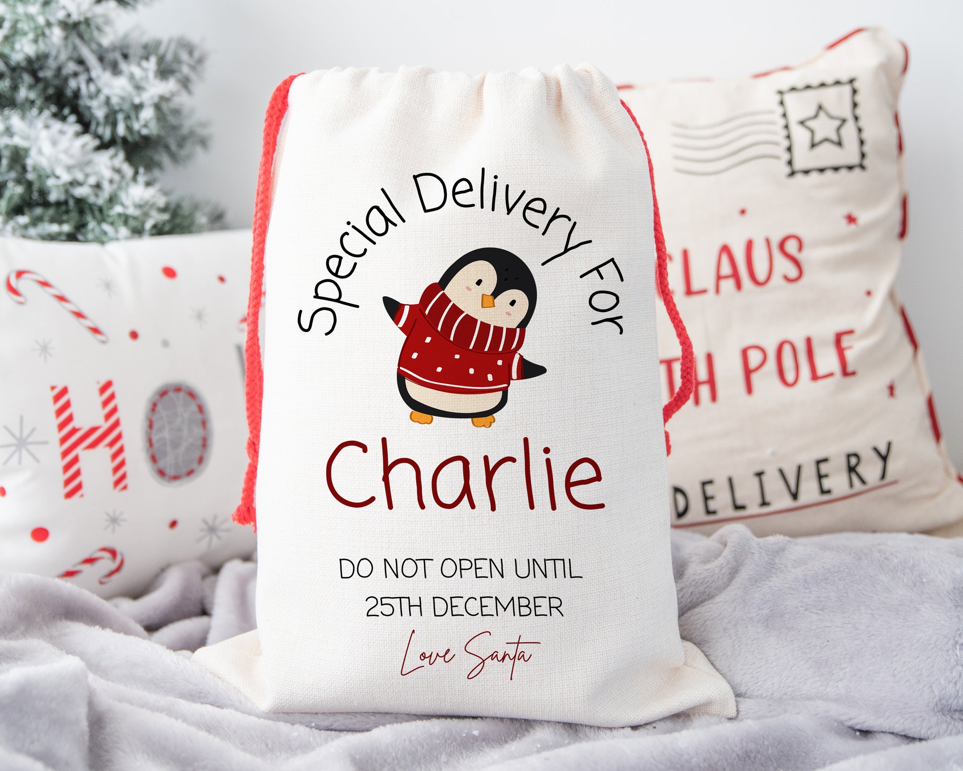 Cute personalised Christmas gift sack with penguin in red scarf – custom “Special Delivery for Charlie” linen Santa sack with red drawstring – festive background with holiday cushions