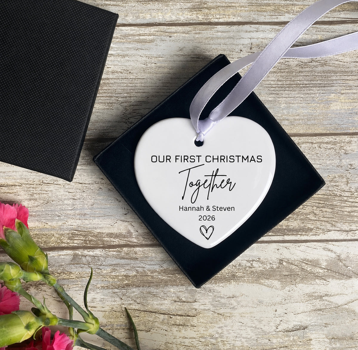 A personalized white heart Christmas ornament with black text reading 'OUR FIRST CHRISTMAS Together Hannah & Steven 2026' and a small heart, hanging from a lavender ribbon over a black square gift box on a rustic wood surface with flowers at the corner.
