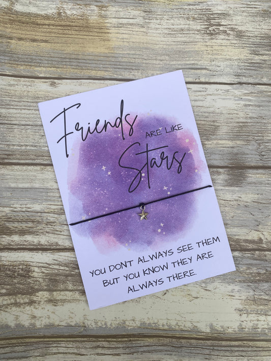 Purple watercolor-themed friendship card with the quote “Friends are like stars” in a handwritten font. A dainty black cord bracelet with a gold star charm is tied across the center, resting on a rustic wooden surface.