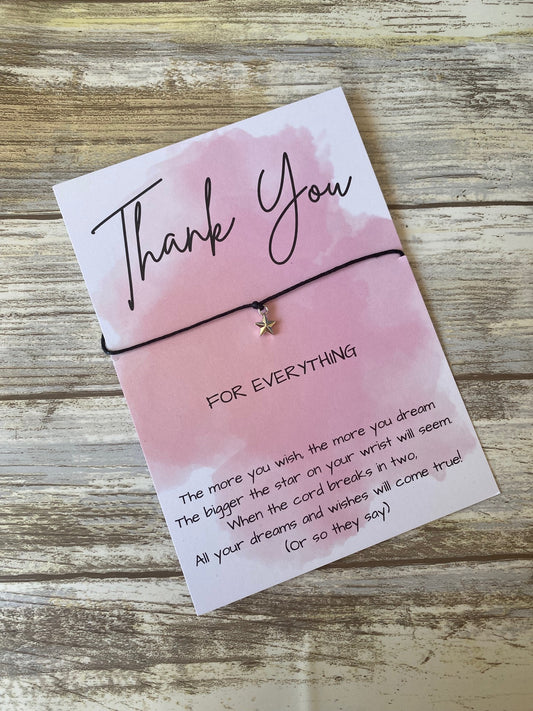 A pink watercolor thank you card featuring a black string bracelet with a small silver star charm. The card reads: “Thank You for everything” with a wish poem printed at the bottom, placed on a rustic wooden surface.