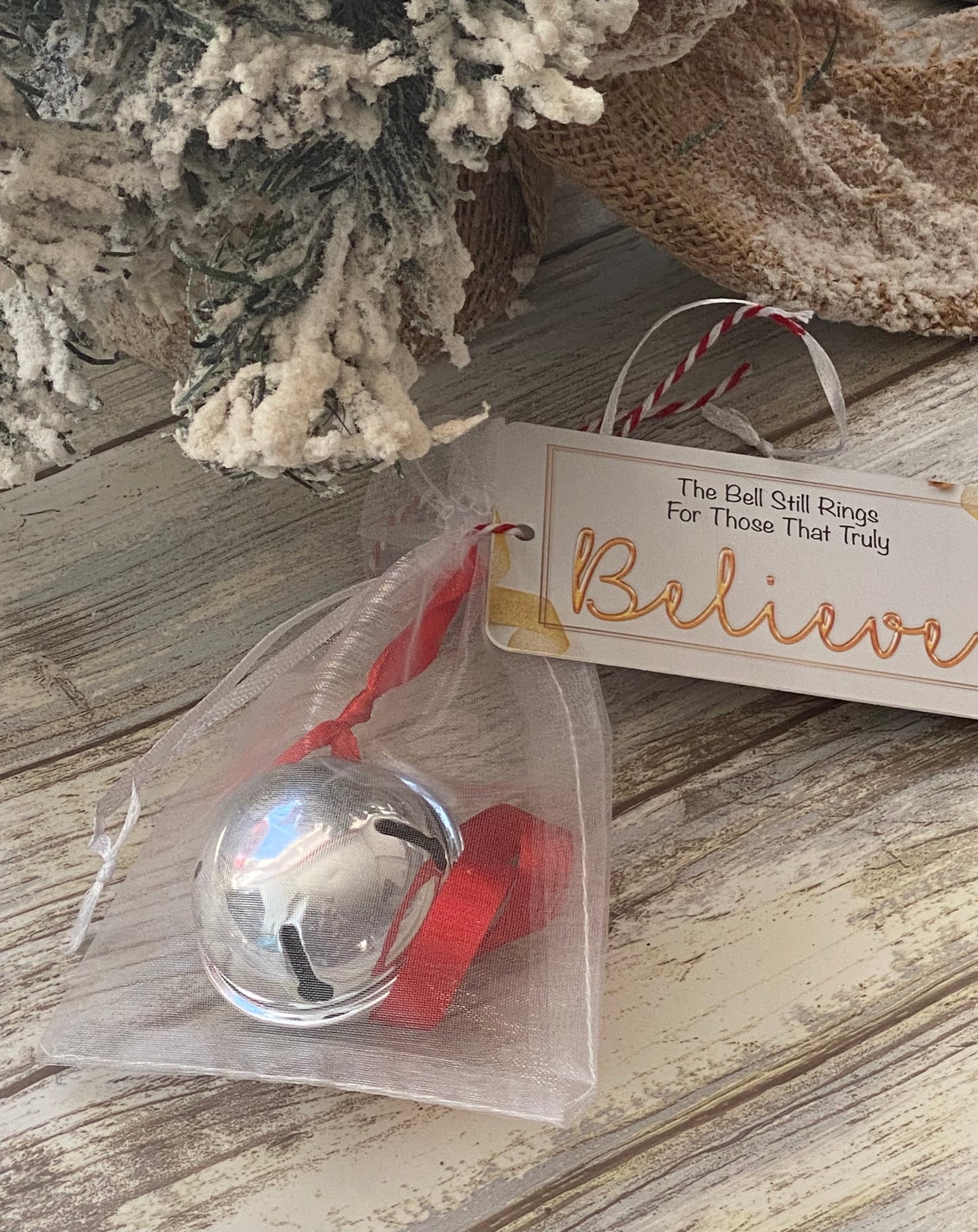 A silver Christmas bell with a red ribbon, placed on a wooden surface, with a decorative rhyming poem card next to it.