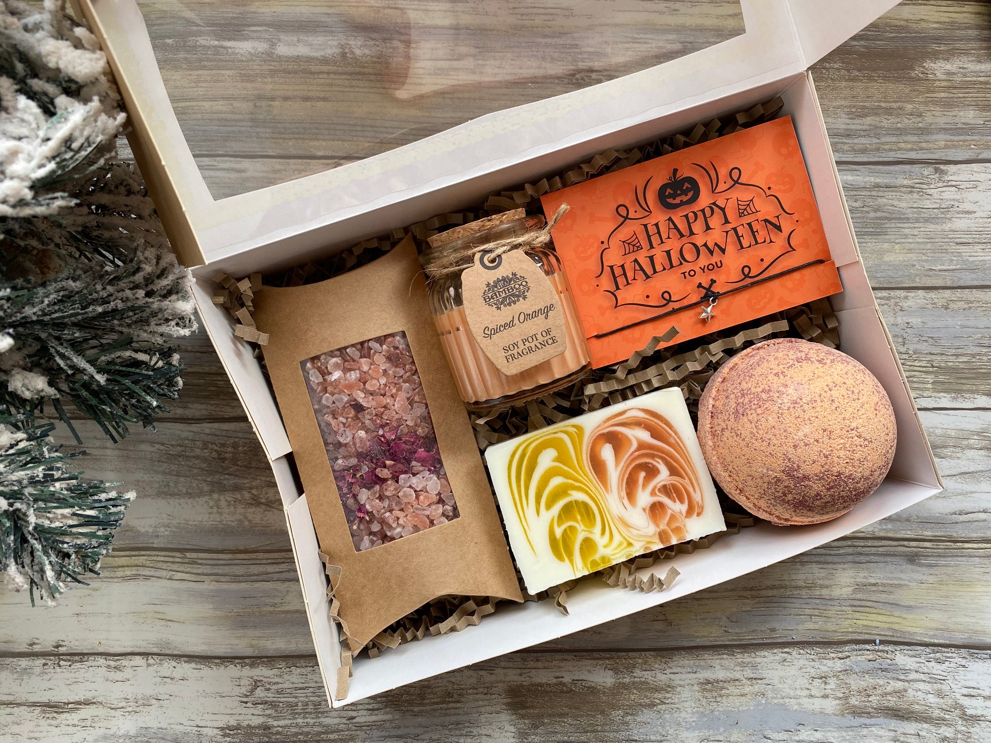 An open gift box displaying a chocolate and orange bath bomb, spiced orange candle, orange and ginger handmade soap bar, and a box of Himalayan salts and rose petals, all arranged on a wooden surface with decorative foliage.