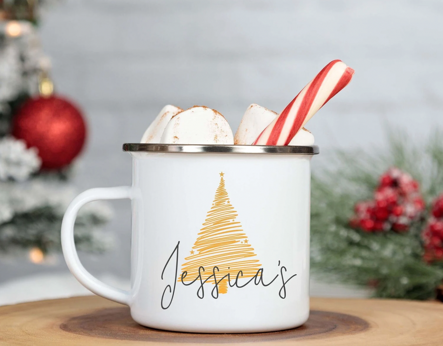 A white enamel mug with a personalised name, silver rim finish, and decorated with a red and white striped candy cane and a Christmas tree graphic. The mug is presented in a festive setting with Christmas decorations in the background.