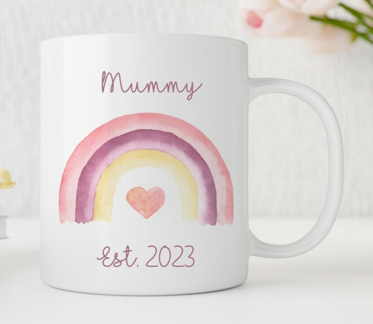 A personalised white ceramic mug with a colourful rainbow and heart design, and the words 'Mummy Est. 2023' printed on it.