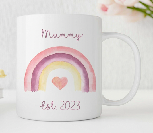 A personalised white ceramic mug with a colourful rainbow and heart design, and the words 'Mummy Est. 2023' printed on it.