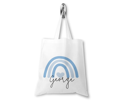 White tote bag hanging on a display hook, featuring a minimalist blue rainbow with a heart in the centre and the name "George" written in black script font underneath.
