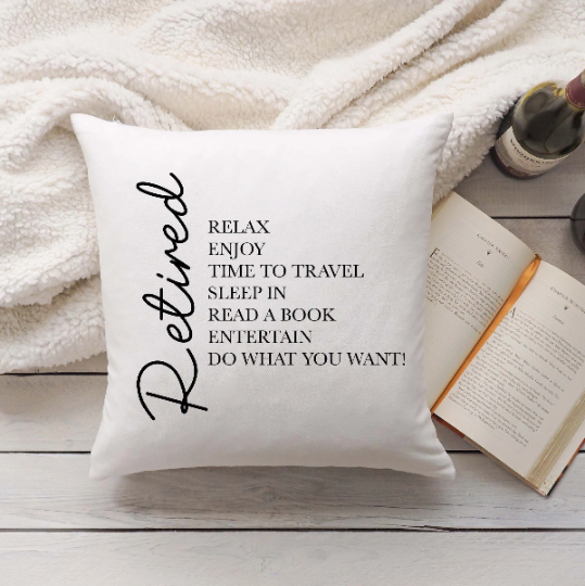 A cushion with black text that reads 'RETIREMENT CELEBRATION CUSHION' with a pen and a book in the background.