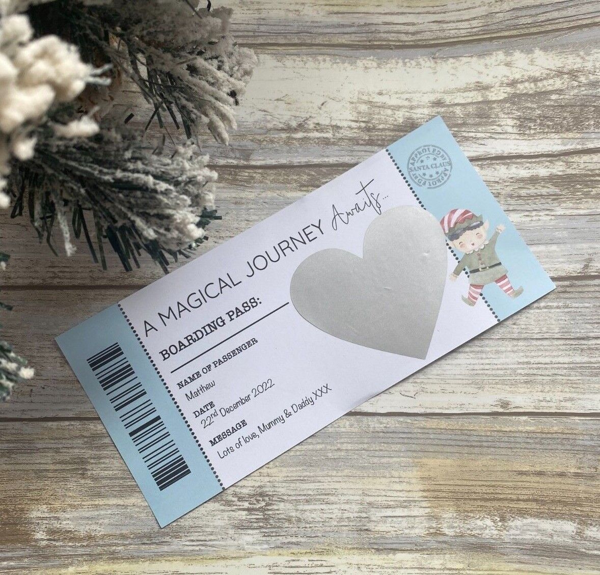 A boarding pass invitation to Lapland with a heart shape on the scratch panel, presented on a wooden surface with Christmas decorations in the background.