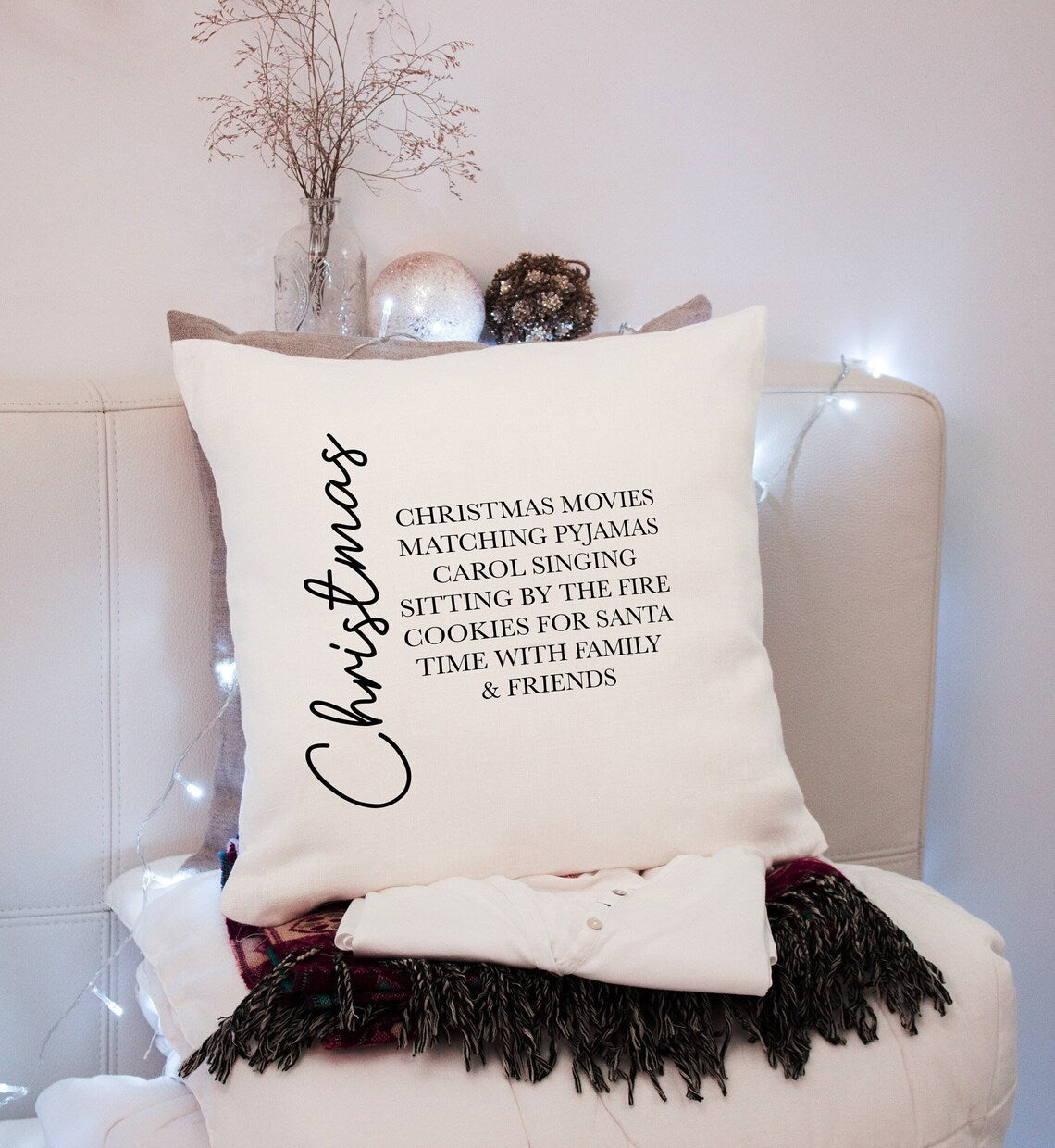 A Christmas-themed cushion with text printed on it, placed on a chair with a decorative branch and lights in the background.
