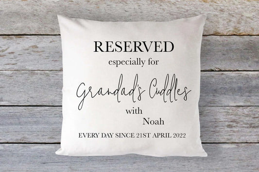 A square-shaped cushion with text that personalises it with a grandchild's name and a date, placed on a wooden surface.