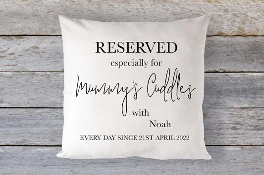 A square-shaped cushion with the text 'RESERVED especially for Mummy's Cuddles with Noah EVERY DAY SINCE 21ST APRIL 2022' printed on it, placed on a wooden surface.