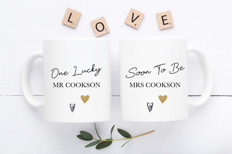 Two white ceramic mugs with printed text and hearts, one labeled 'One Lucky MR COOKSON' and the other ' Soon To Be MRS COOKSON', presented on a white surface with the letters 'LOVE' arranged at the top and a small branch to the side.