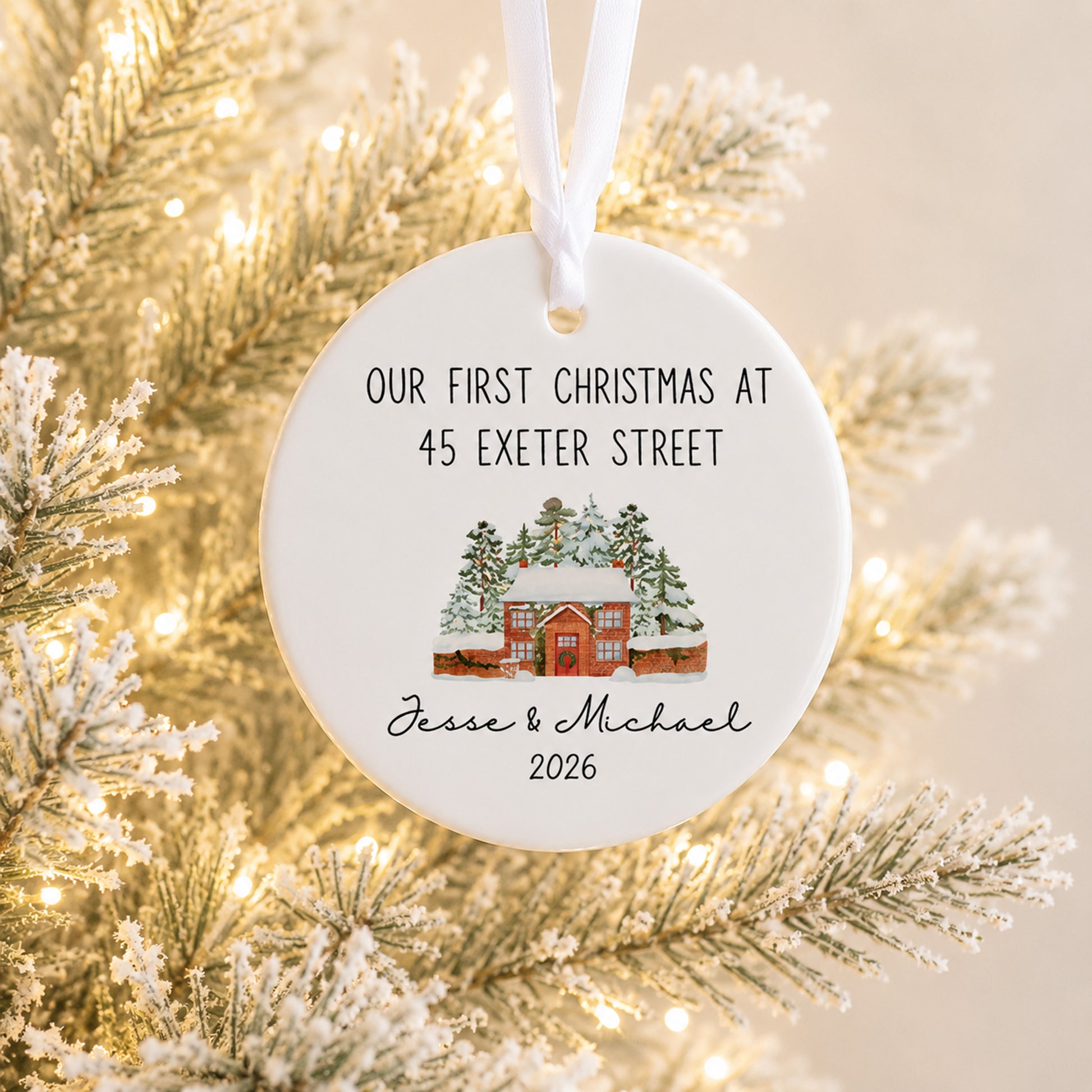 A round white ceramic Christmas ornament hanging from a white ribbon, printed with the text 'OUR FIRST CHRISTMAS AT 45 EXETER STREET' and 'Jesse & Michael 2026' above a small illustrated cabin scene. The ornament is photographed in front of blurred flocked pine branches with warm string lights.