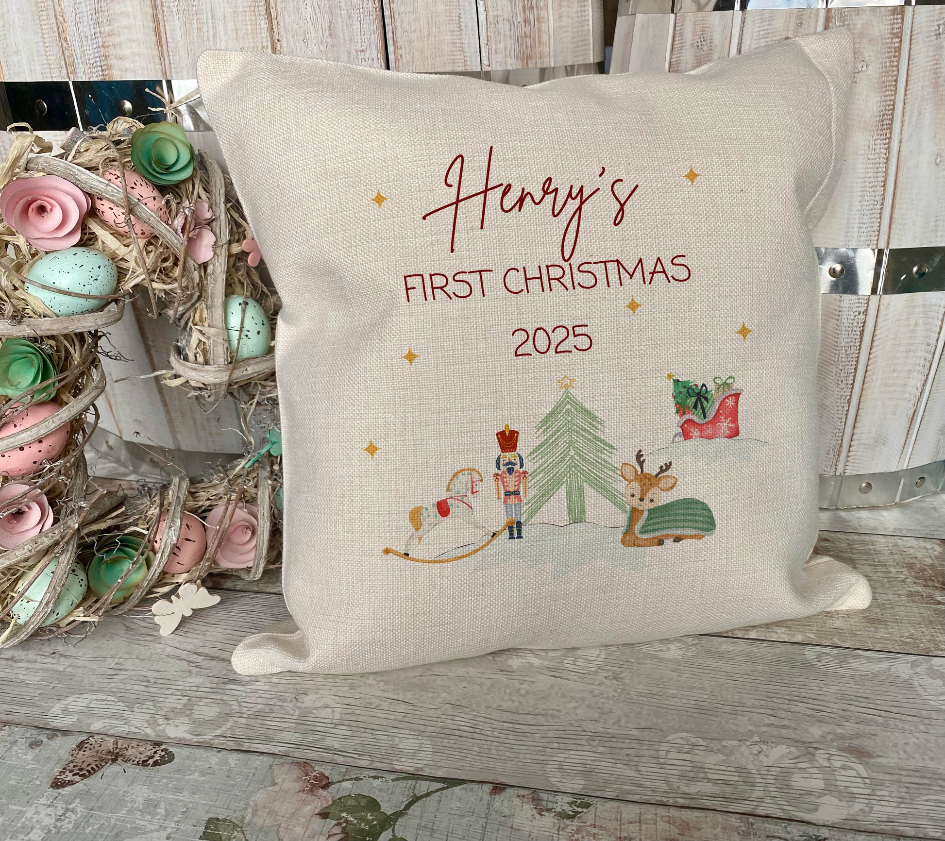 Decorative pillow with 'Henry's First Christmas 2025' text and festive illustrations on a wooden surface.