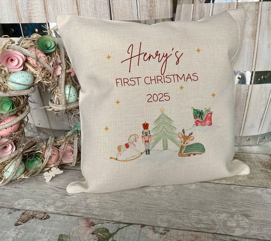 Decorative pillow with 'Henry's First Christmas 2025' text and festive illustrations on a wooden surface.