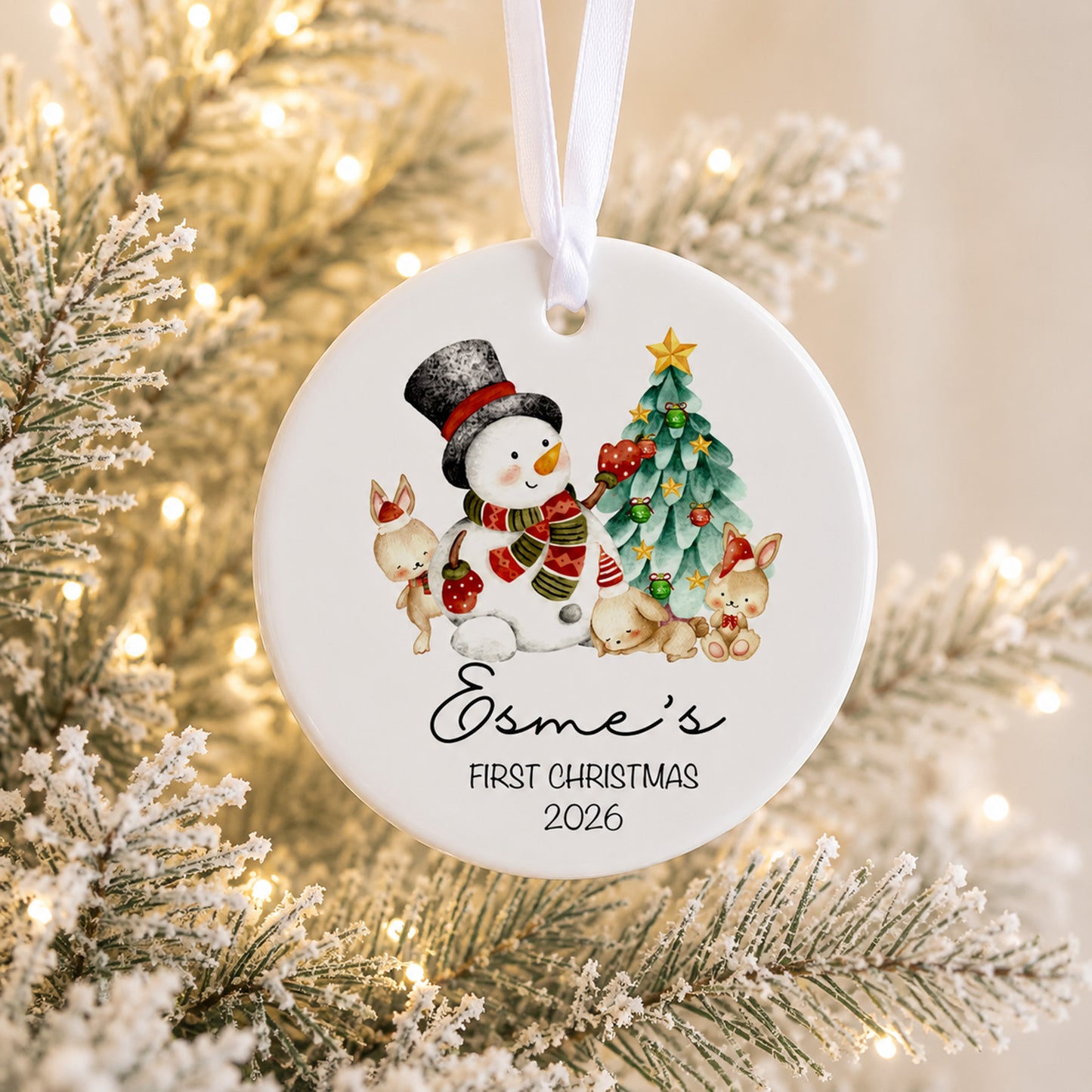 Personalized round white Christmas ornament hanging from a white ribbon, featuring a watercolor snowman, tree, rabbits, and the text 'Osme's FIRST CHRISTMAS 2026' against a blurred festive tree background with warm lights.
