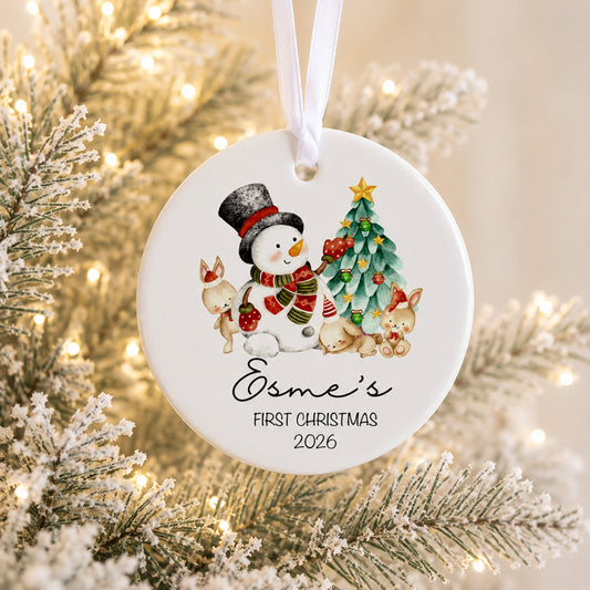 Personalized round white Christmas ornament hanging from a white ribbon, featuring a watercolor snowman, tree, rabbits, and the text 'Osme's FIRST CHRISTMAS 2026' against a blurred festive tree background with warm lights.