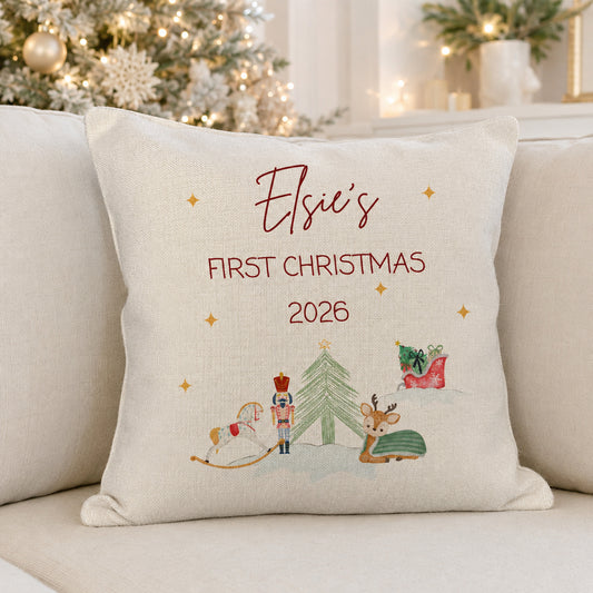 A personalized cream-colored throw pillow with embroidered text reading 'Elsie's FIRST CHRISTMAS 2026' and a festive Christmas illustration, resting on a light beige sofa with a blurred Christmas tree in the background.