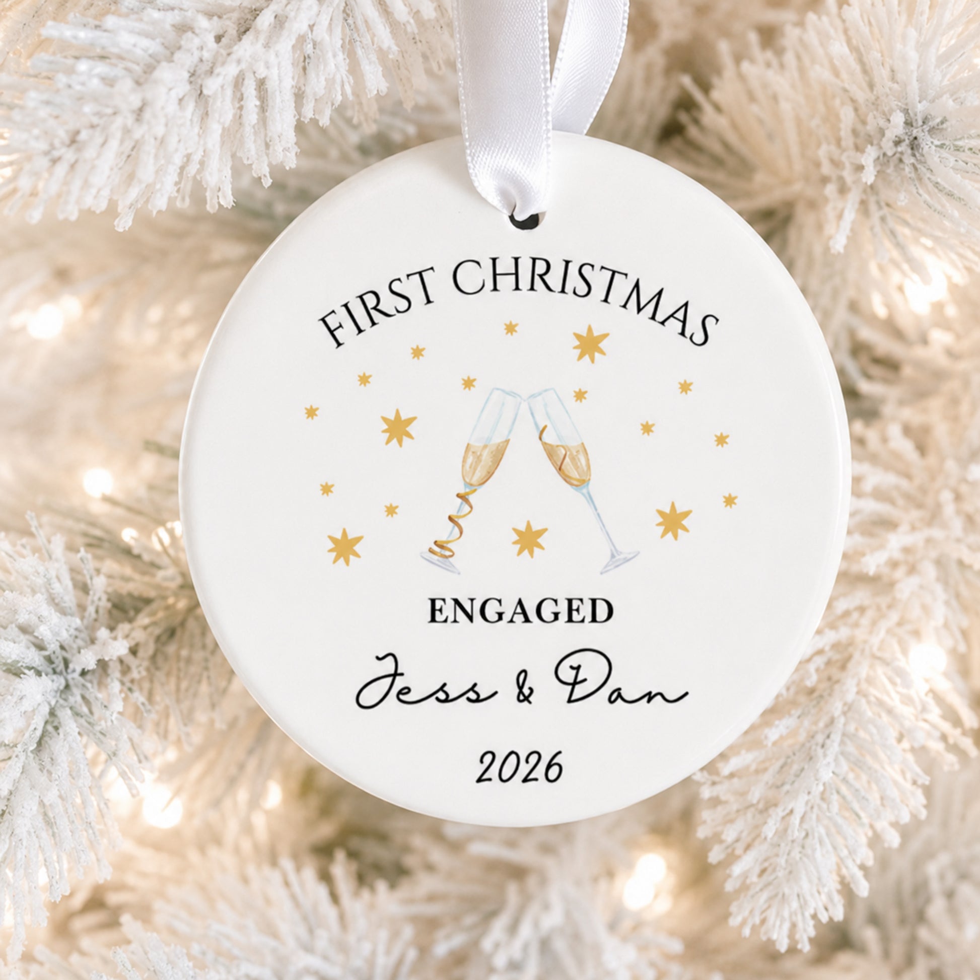A round white personalized Christmas ornament hanging from a white ribbon, printed with 'FIRST CHRISTMAS ENGAGED Jess & Dan 2026' and a champagne toast illustration, shown against frosted tree branches with warm lights.