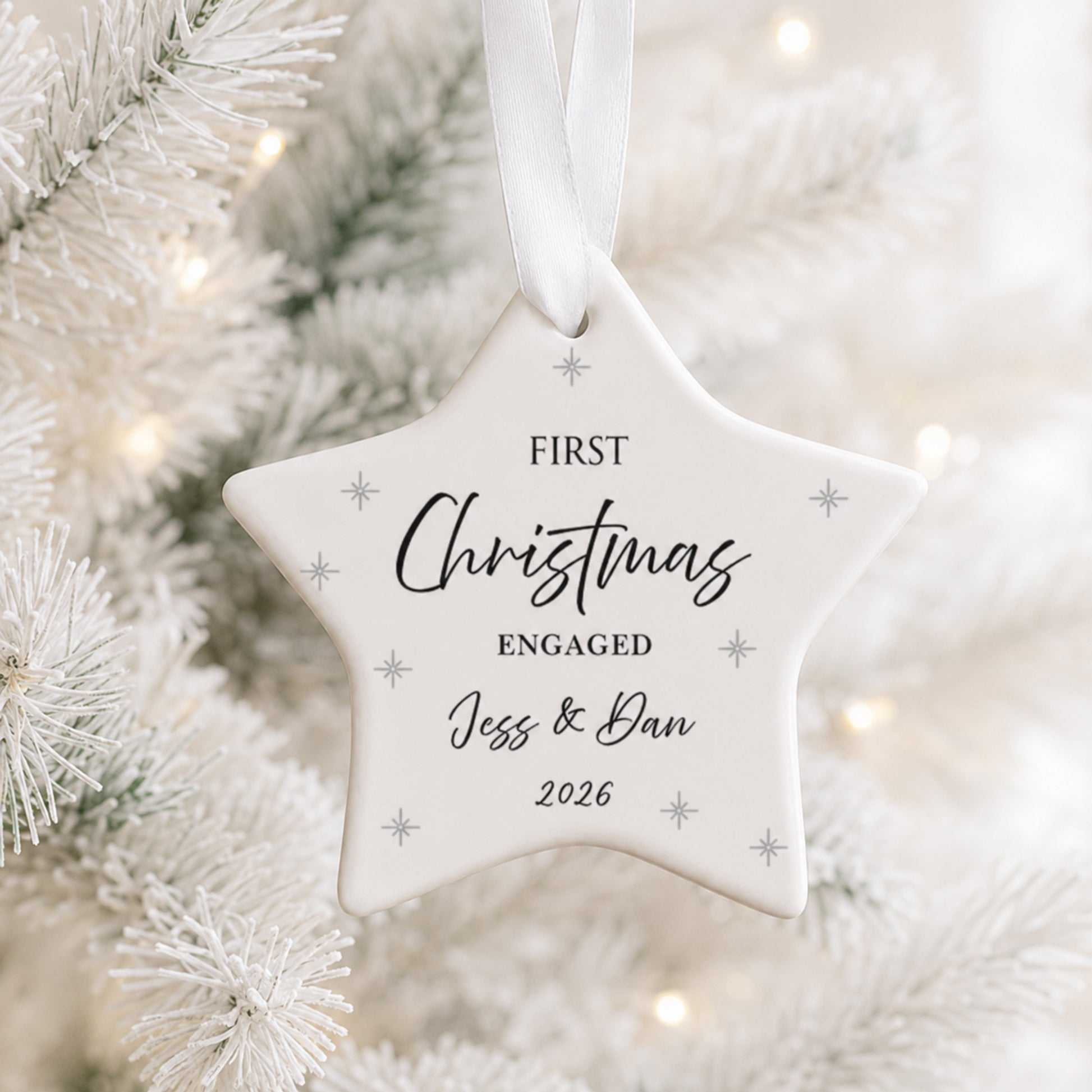 A white star-shaped Christmas ornament hanging from a white ribbon, printed with the words 'FIRST Christmas ENGAGED Jess & Dan 2026' and small silver snowflake motifs, set against a blurred white Christmas tree background with warm lights.