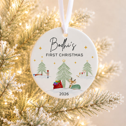 A personalized round ceramic Christmas ornament hanging from a white ribbon, featuring the text 'Bodhi's FIRST CHRISTMAS 2026' and a festive winter scene with trees, a Santa Claus figure, and a reindeer, set against a blurred golden Christmas tree background.