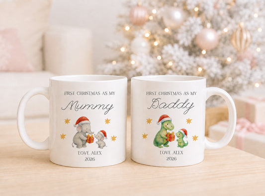 Two white First Christmas as my Mummy and Daddy ceramic mugs with personalized Christmas designs, displayed side-by-side on a light wooden table with a blurred festive background of a decorated Christmas tree.