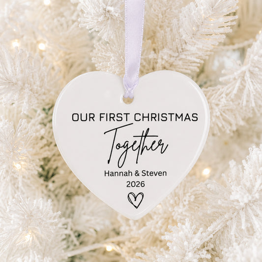 A white heart-shaped personalized Christmas ornament hanging from a white frosted tree with warm string lights. The ornament is printed with the text 'OUR FIRST CHRISTMAS Together Hannah & Steven 2026' in black lettering.