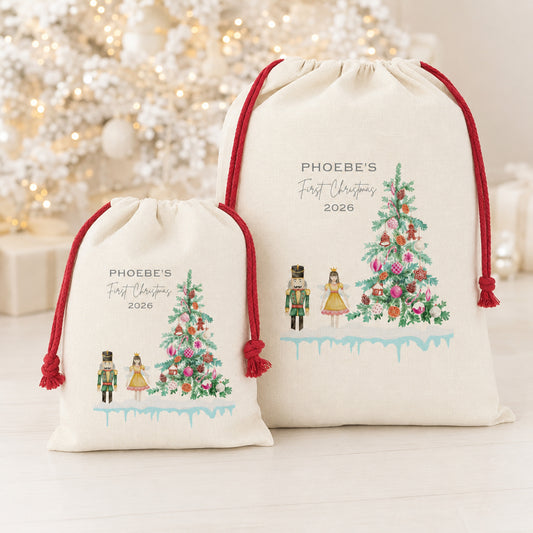 Two personalized cream-colored drawstring gift bags with red cords, featuring a watercolor Christmas tree and nutcracker design, displayed in front of a blurred festive background.