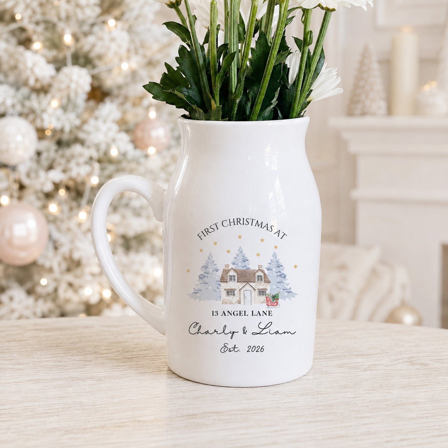 A white ceramic flower jug with a handle, decorated with a personalized Christmas illustration and text reading 'FIRST CHRISTMAS AT 13 ANGEL LANE Charly & Liam Est. 2026', filled with green stems and white flowers, set on a light wooden surface with a blurred festive Christmas tree in the background.