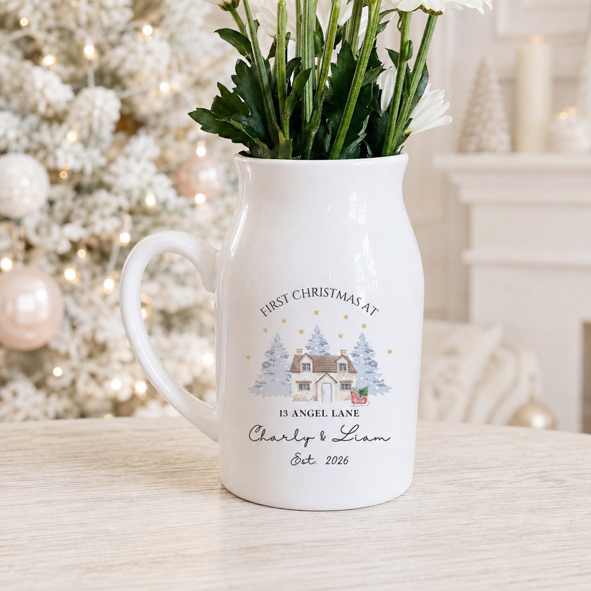 A white ceramic flower jug with a handle, decorated with a personalized Christmas illustration and text reading 'FIRST CHRISTMAS AT 13 ANGEL LANE Charly & Liam Est. 2026', filled with green stems and white flowers, set on a light wooden surface with a blurred festive Christmas tree in the background.