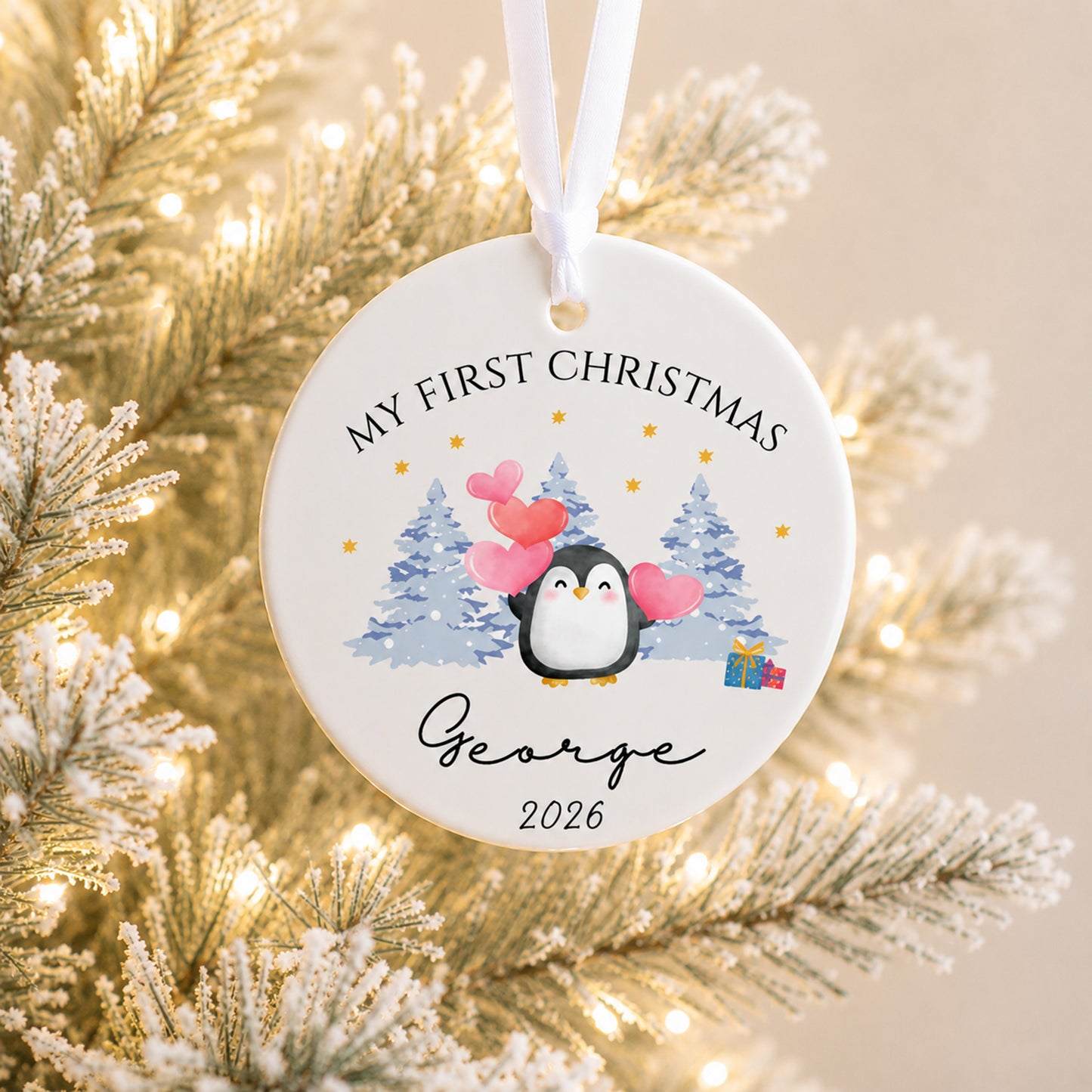 A personalized round ceramic Christmas ornament hanging from a white ribbon, featuring a penguin illustration with pink hearts, blue pine trees, and the text 'MY FIRST CHRISTMAS George 2026'. The ornament is photographed against a softly blurred festive background of frosted branches and warm string lights.