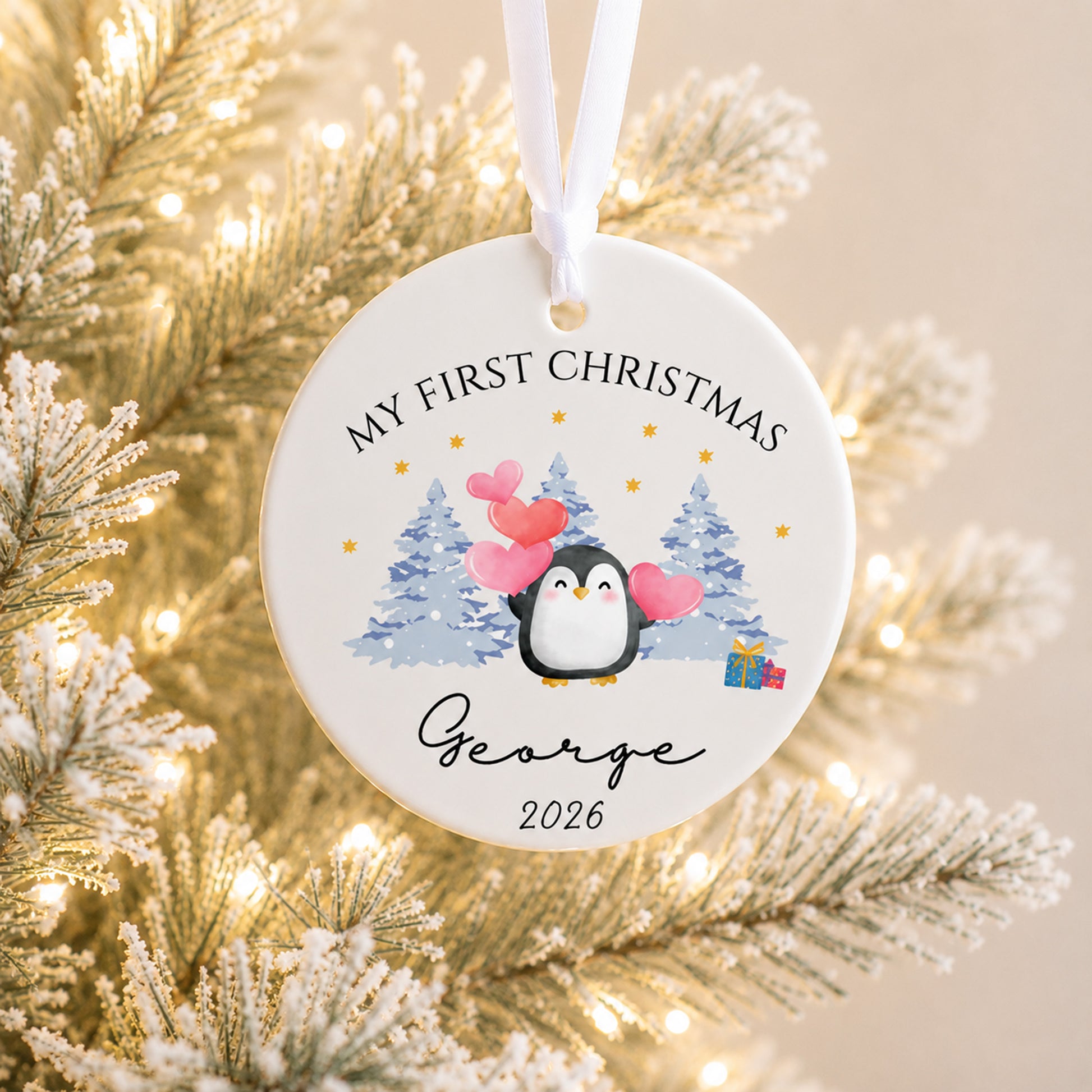 A personalized round ceramic Christmas ornament hanging from a white ribbon, featuring a penguin illustration with pink hearts, blue pine trees, and the text 'MY FIRST CHRISTMAS George 2026'. The ornament is photographed against a softly blurred festive background of frosted branches and warm string lights.
