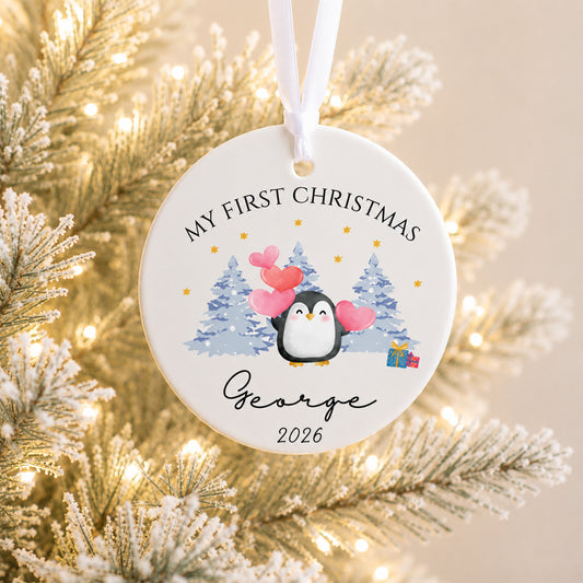 A personalized round ceramic Christmas ornament hanging from a white ribbon, featuring a penguin illustration with pink hearts, blue pine trees, and the text 'MY FIRST CHRISTMAS George 2026'. The ornament is photographed against a softly blurred festive background of frosted branches and warm string lights.