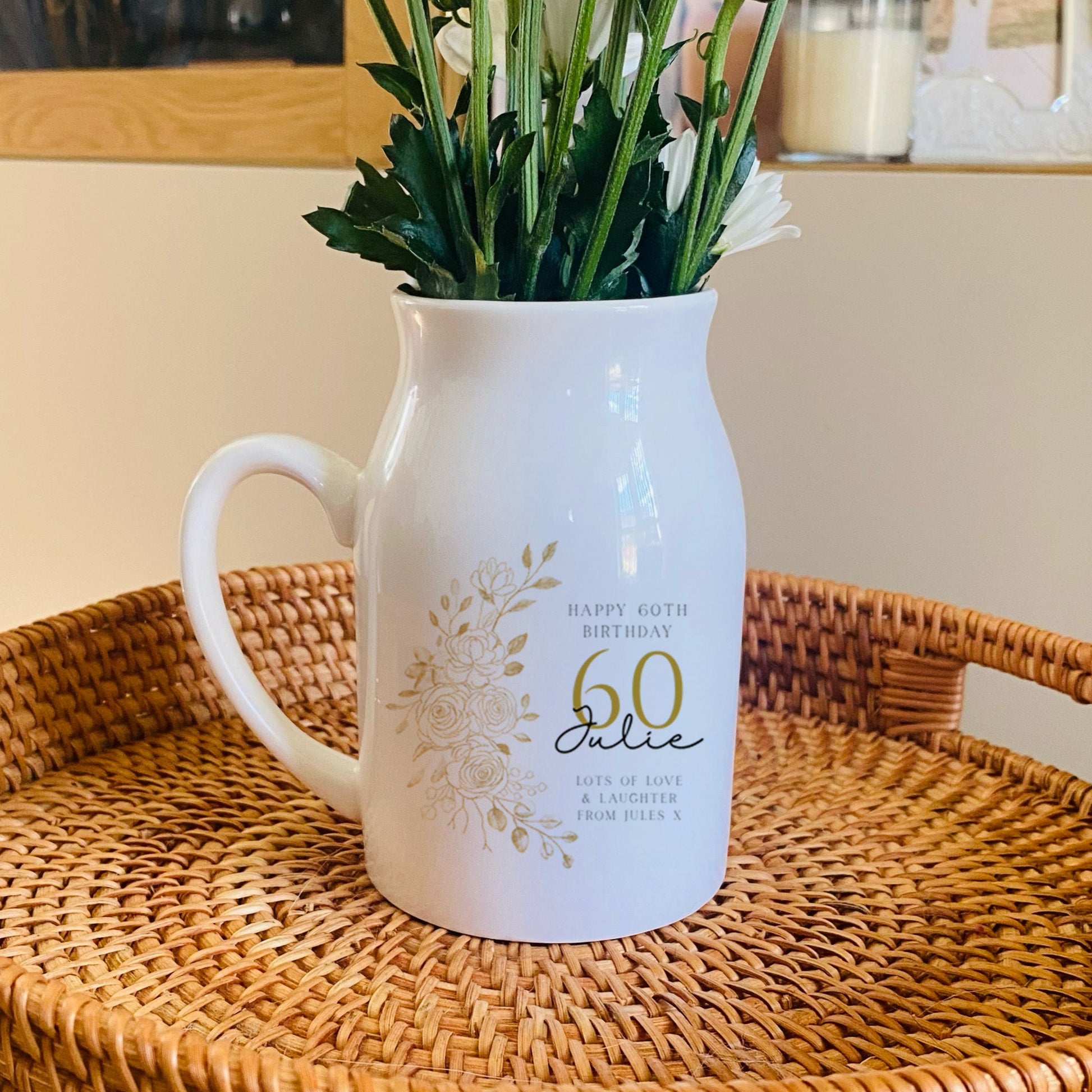 White 60th birthday vase pitcher with floral arrangement on a woven tray in a home setting