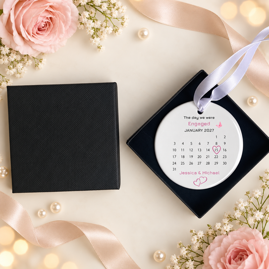 A personalized engagement ornament displayed next to its black presentation box on a styled flat-lay background with pink roses, baby's breath, pearls, and satin ribbons.